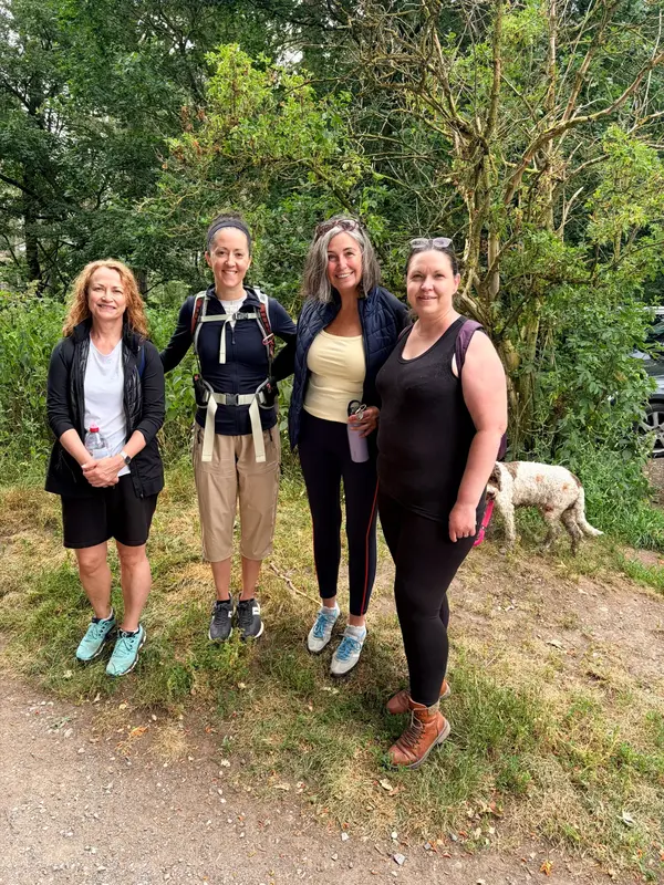 Group of four smiling midlife women in walking gear in the forest, with a dog in the background